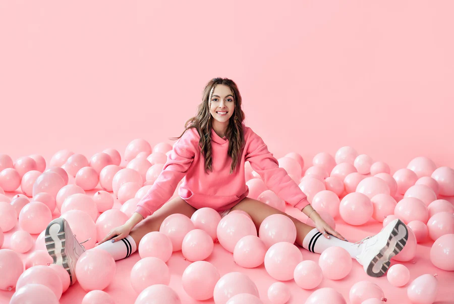Mladá žena sedí na zemi obklopená ružovými balónmi, usmieva sa a nosí pohodlný outfit.
