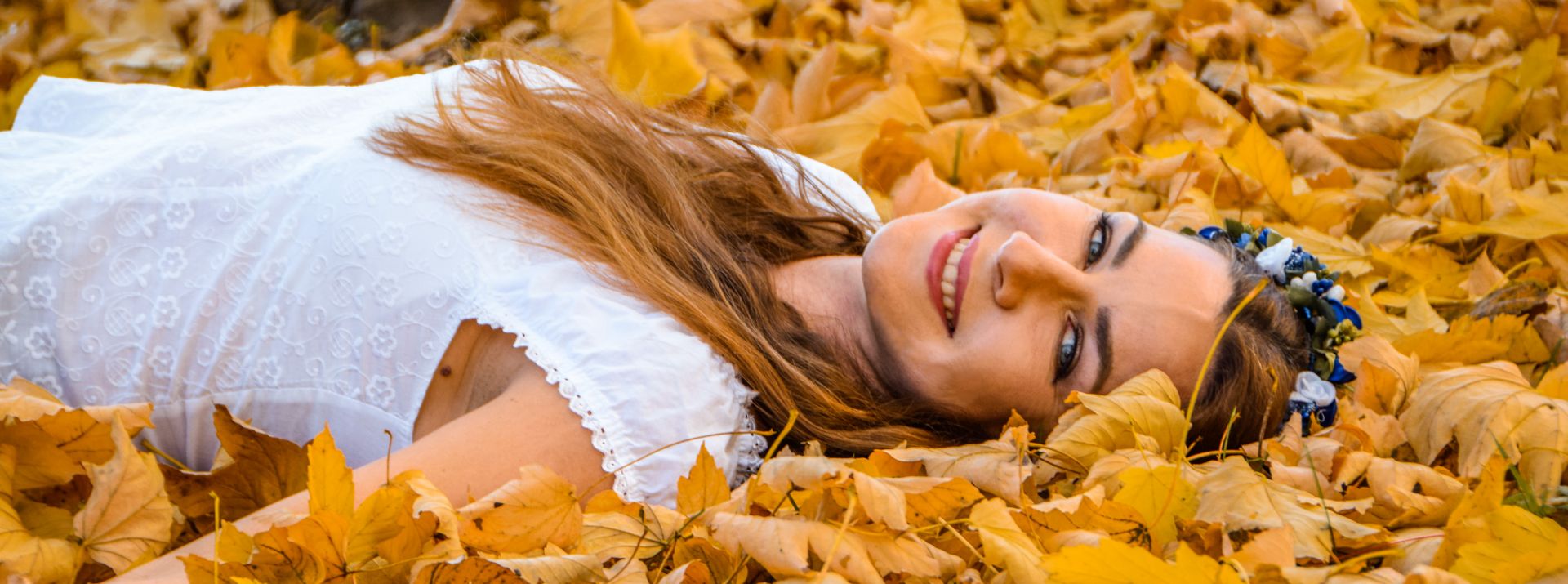 Žena leží na zemi obklopená žltými listami, usmieva sa a má kvetinový veniec.
